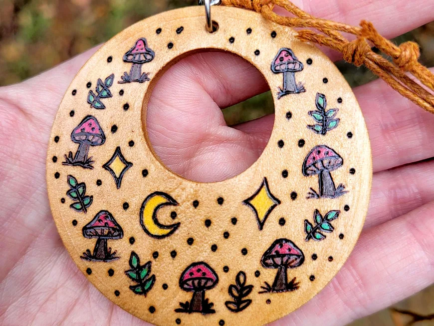 A round wood pendant burned with mushrooms, moons and leaves, on a cord
