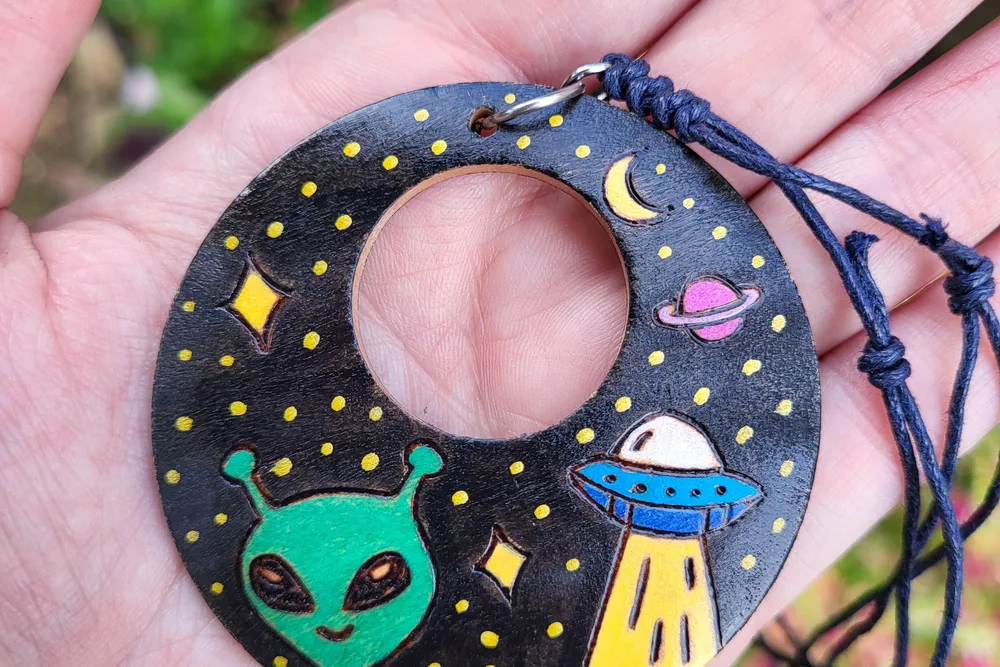 A hand holding a round wood pendant painted with a green alien, a UFO and a starry night sky